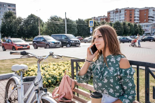 Hermosa chica sentada en el banco, bicicleta y bolsa. En verano al aire libre. Estilo de vida en la ciudad. Llama por teléfono, en su mano una taza de café. El concepto de comunicación en línea en las redes sociales . — Foto de Stock