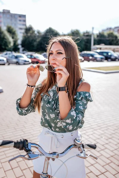 Hermosa chica se para con una bicicleta en verano en la ciudad. Lleva gafas de sol. En verano al aire libre. Estilo de vida en la ciudad . — Foto de Stock
