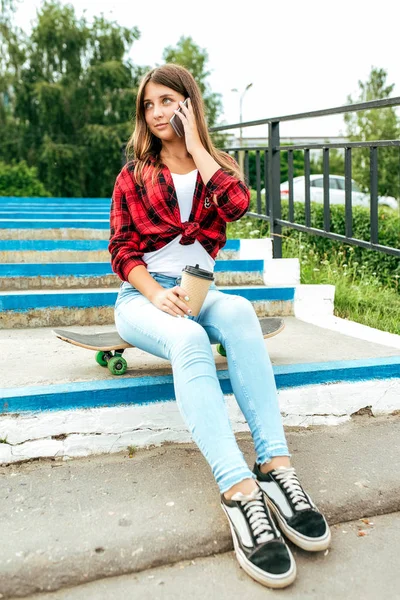 Gadis remaja duduk di skateboard, berbicara di telepon, memegang cangkir dengan teh di tangannya. Di musim panas di kota di alam . — Stok Foto
