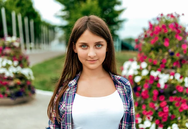 Dívčí studentka na košili v létě v parku stojí na pozadí květin. Šťastný úsměv, odpočívající po škole. Pokožka s dlouhými vlasy. — Stock fotografie