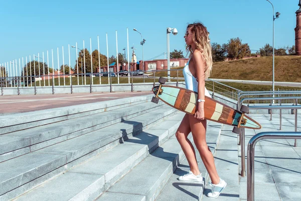 Gelooid meisje in een wit lichaam, lopen de trap in de zomer in de stad. In de hand van een skateboard, Longboard. Lang haar gebruinde huid. Vrije ruimte voor tekst. — Stockfoto