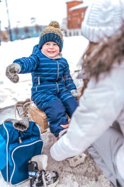 Genç anne kış aylarında halka açık buz pateni pistine paten koyar. Küçük mutlu çocuk gülümseyerek elini yana doğru gösteriyor, mavi tulum içinde, eldivenler karda. Kışın buzda buz pateni yapmayı öğrenmek.