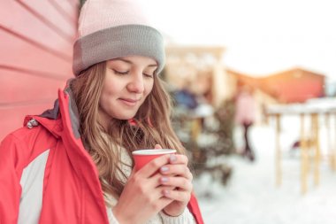 Güzel kız öğrenci, kış tatili, kar arka planda Yeni Yıl tatili. Mutlu gülümseyerek, elinde sıcak bir içecek, kahve çayı ile bir fincan tutarak. Isıtıyor ve hayaller kuruyor. Metin için boş alan.