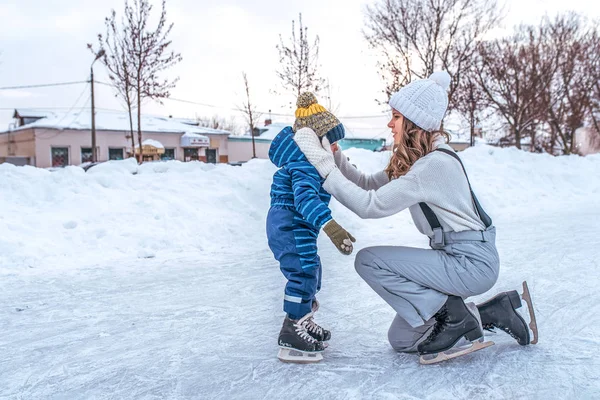 Seorang ibu muda meluruskan anak-anaknya cap boy. Di musim dingin di kota di gelanggang es. Dalam seluncur pakaian musim dingin, di latar belakang salju. Ruang kosong untuk teks . — Stok Foto