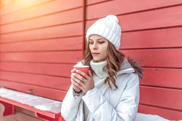 Piękna dziewczyna zimą w mieście na tle czerwonych okiennic. W ręku trzyma szklankę z ciepłym napojem herbaty lub kawy, nagrzewa się w weekend podczas wakacji. Wolne miejsce na tekst. — Zdjęcie stockowe