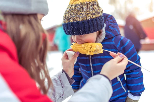 Мальчик зимой в теплой синей куртке. Он ест вареную кукурузу, отдыхает на праздники в новогодние праздники. Концепция ухода за детьми в холодное время года . — стоковое фото