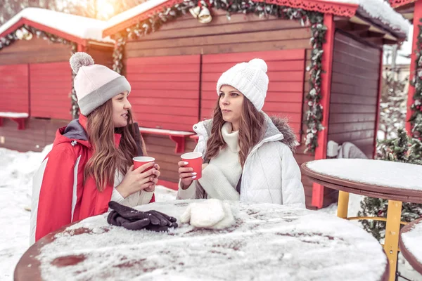 Dua gadis membuat teman-teman di musim dingin di meja tertutup salju, berbicara dan mengobrol. Di tangan cangkir teh kopi, sarapan camilan di alam pada hari libur dan liburan Tahun Baru . — Stok Foto