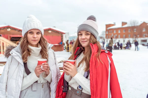 Two girls make friends in winter pose. In hands of cups coffee tea, snack breakfast on nature in New Year holidays and vacations. Keep warm on a hot drink. Emotions of joy, smiles and fun at resort.