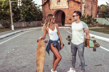 Şehirde yaz aylarında ayakta duran genç bir çift, kaykaylar elinde, longboard. Hafta sonları iletişim, gençlerin yaşam tarzı. Mutlu gülümsüyor konuşmak. Sevinç eğlenceli gülümseme Duyguları.