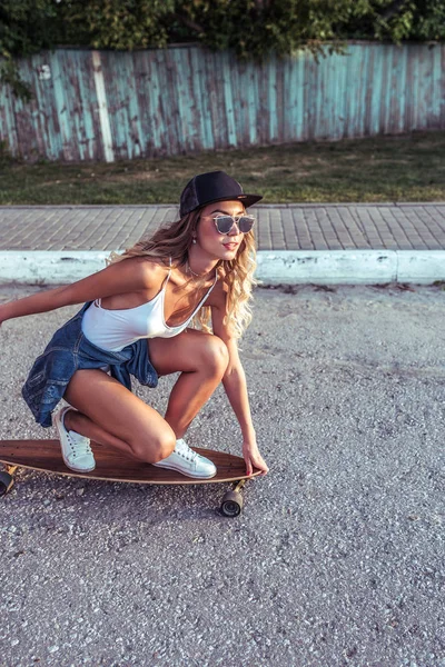 Gadis di skateboard, di musim panas di kota, tubuh putih, jaket, jins, topi baseball hitam, kacamata hitam. Di latar belakang jalan dan aspal. Rides beristirahat di akhir pekan . — Stok Foto