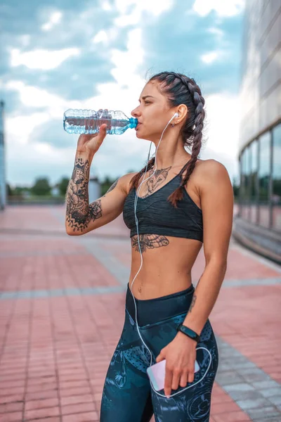 Mujer atleta en tatuajes, ciudad de verano, descansando después del ejercicio físico. Top de ropa deportiva. El concepto de fuerza, feminidad, coraje, confianza, inspiración, motivación. Bebe agua de una botella . — Foto de Stock