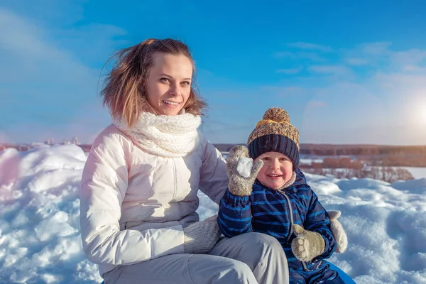 Seorang ibu muda dengan anak laki-laki berusia 4 tahun. Duduk di musim dingin melawan salju dan langit. Bermain di luar ruangan yang menyenangkan di liburan akhir pekan . — Stok Foto