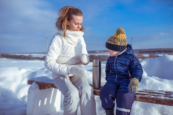 Молодая мать с маленьким сыном, мальчик 4 лет. Зимой сидит на скамейке на фоне снега и заносов. Термос с горячим чаем и супом. Кемпинг в зимние каникулы с ребенком . — стоковое фото