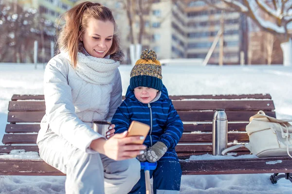 Молодая женщина с маленьким сыном 4 лет. Разговариваю по видеосвязи. Зимой сидит на скамейке на фоне снега и заносов. Онлайн общение в Интернете, фотографируйте себя . — стоковое фото