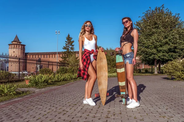 Dvě přítelkyně, krásné dívky jsou v létě ve městě. Skateboard, dlouhý prkno. Vyčiněná kůže má dlouhé vlasy. Koncept módního životního stylu mladých lidí. Volné místo pro text. — Stock fotografie