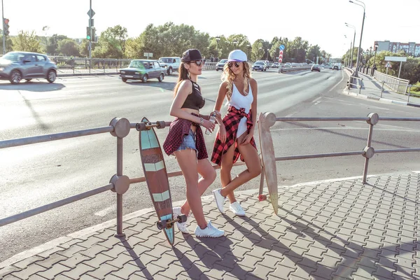 Zwei Freundinnen, schöne Mädchen stehen Sommerstadt, Hintergrundstraße, Kreuzung. Leute, die lächeln. Skateboard, Longboard. gebräunte Haut hat lange Haare. das Konzept Mode Lifestyle junge Leute. — Stockfoto