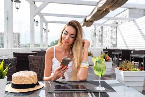 Schöne Sommermädchen im Café, hört Musik mit Kopfhörern. Auf dem Tisch liegt ein Cocktail, eine Tablette. Mobiltelefon, Online-Anwendung soziale Netzwerke. glücklich lächelnd in einem Restaurant ausruhen. — Stockfoto