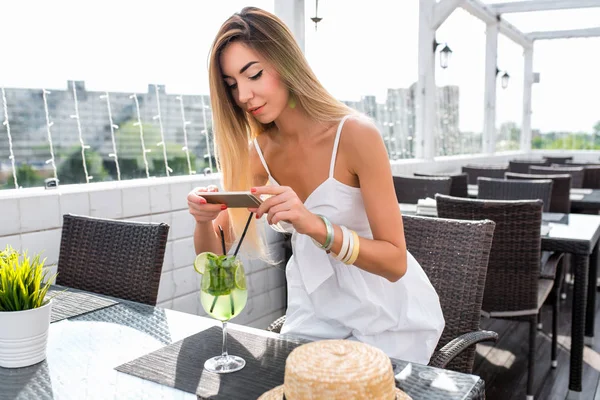 Restoran elbisesi giymiş güzel kadın, kız yaz kafesi. Fotoğraf kokteyli masada, şapka. Uzun saç günlük makyaj. İşten sonra dinlen. Fotoğraf sosyal ağlar kavramı, video kayıt akıllı telefon.