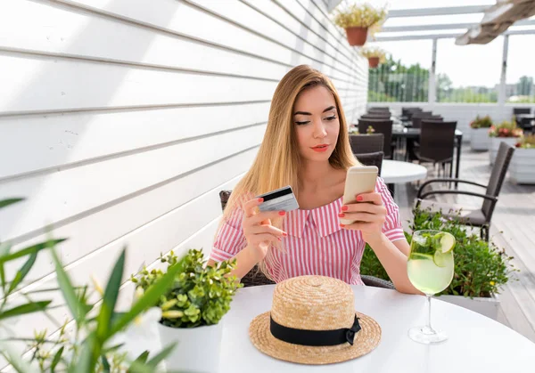 Hermosa mujer en un vestido de restaurante, chica en la cafetería de verano. Concepto de comprar y pagar la cena con tarjeta de crédito. En la mano del teléfono hay una tarjeta de plástico. Banca en línea, solicitud en línea . —  Fotos de Stock