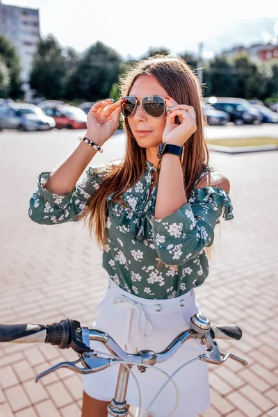 La muchacha en verano en la ciudad se para con la bicicleta en la ciudad. Lleva gafas de sol. Blusa verde y pantalones cortos de falda blanca. Estilo de vida activo el fin de semana. Actividades exteriores . — Foto de Stock