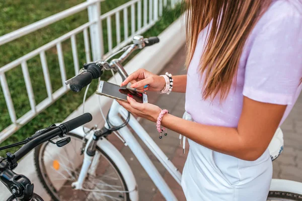 Женщина летом стоит на велосипеде, активирует приложение, покупая велосипед напрокат на парковке, в руках телефона. Онлайн выбор маршрута на карте города. Велоспорт в выходные. Карта маршрута по телефону . — стоковое фото