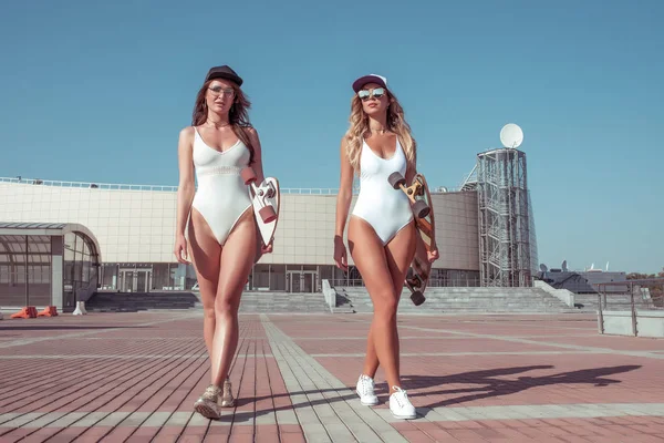 İki kız kadın beyaz mayo baud, yaz şehir sokakta gider. Uzun tahta kaykay güneş gözlüğü. Yürüyen kızlar. Konsept tarzı moda, yaşam tarzı gençler. Uzun saç tabaklanmış şekil.