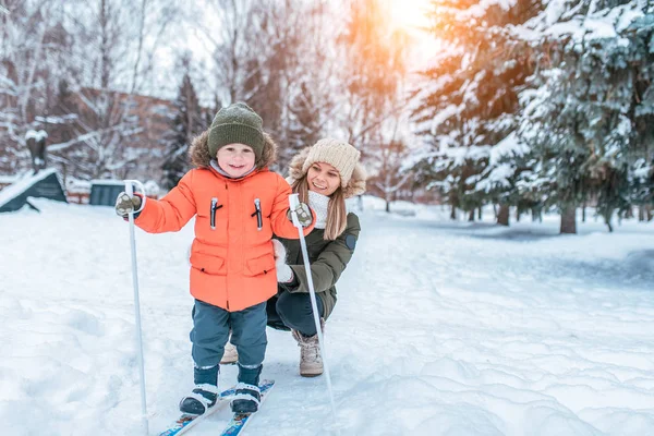 Young mother woman happy smiling, son boy 3 years old laughs. In winter, outside park, background is snow drifts Christmas tree. Free space. Childrens skis, skiing lessons, first steps, child care.