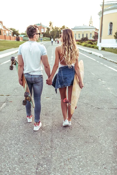 Young couple man woman walk summer city, background road view from rear. In hands skateboard, longboard. Holding hands, concept love, caring, understanding, relationship. Empathy care inspiration.