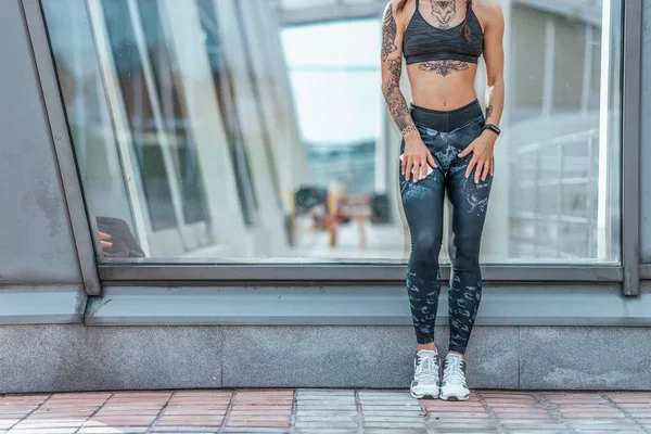 Girl stands glass case, summer city woman fitness sport, active lifestyle youth. Motivation power, achievement, success. Free space. Phone tattoos fitness bracelet sportswear, leggings, top sneakers.
