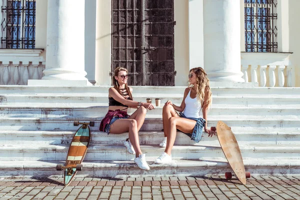 Zwei Freundinnen Mädchen, Frauen Schwestern, sprechende Schritte Sommerstadt, Skateboard, Tassen Kaffee, Tee. Konzept Urlaubswochenende, glückliche Umarmung. das Konzept des Modestils, des modernen Lebensstils. Freiraum. — Stockfoto