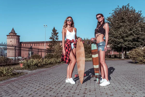 Zwei Freundinnen Mädchen, Frauen Schwestern, posieren im Sommer in der Stadt, Skateboard, lässig tragen kurze Badehosen. das Konzept eines Wochenendausflugs, Modestil, moderner Lebensstil. Freiraum. — Stockfoto