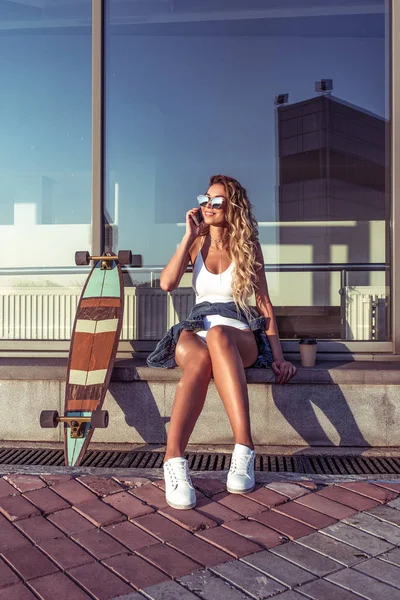 Frau Sommer in der Stadt weißen Körper. Skateboard schöne schlanke, braun gebrannte Figur, Sonnenbrille. Telefon, Online-Bewerbungsgespräche. das Konzept des Modestils, des modernen Lebensstils. — Stockfoto