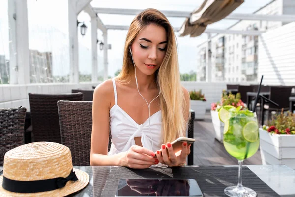 Eine Frau in einem Café sitzt an einem Tisch, in der Hand ein Telefon, Kopfhörer. hört Musik, schreibt eine Nachricht in sozialen Netzwerken. Online-Anwendung im Internet. schöne Mädchen lange Haare. — Stockfoto
