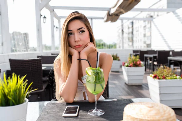 Frau weißes Kleid Sommercafé, traurige Vorfreude, traurig. Unzufrieden, Gedanken an ein negatives Durcheinander. Frühstück Mittagessen, Snack-Restaurant. Warten auf ein Date. Auf dem Tisch liegt ein Telefon. — Stockfoto