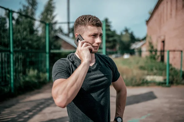 Um homem atleta chama ao telefone, depois de um treino, descansar depois de uma corrida de treino de fitness. Verão na cidade no fundo do campo atlético. Conceito de chamada de aplicação online nas redes sociais . — Fotografia de Stock