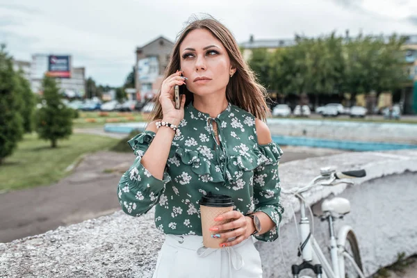 Žena v létě ve městě, telefonuje a v ruce drží šálek kávy. Kolo na pozadí stromů. Obchodní hovor, obchodní holka po práci. Snídaně nebo svačinka u oběda. — Stock fotografie
