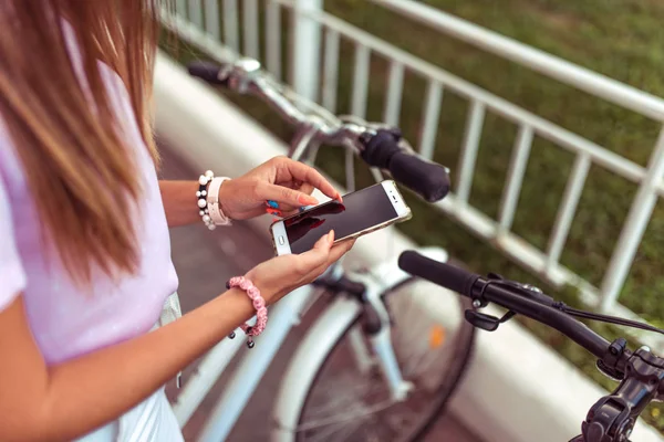 Gadis di musim panas di kota dengan telepon genggam, pilihan rute, latar belakang sepeda parkir. Aplikasi online untuk menyewa sepeda. Tangan wanita cerdas manikur gelang dan rambut panjang . — Stok Foto