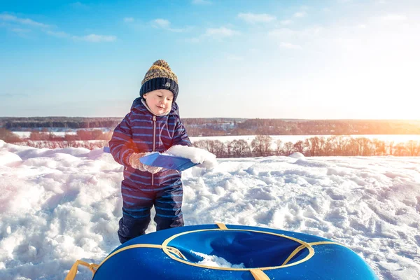 Ein kleiner Junge spielt im Winter draußen mit Schnee, einem Paddel aus aufblasbaren Schläuchen zum Skifahren. warme Winterkleidung und Hut. Freiraum für Text. — Stockfoto