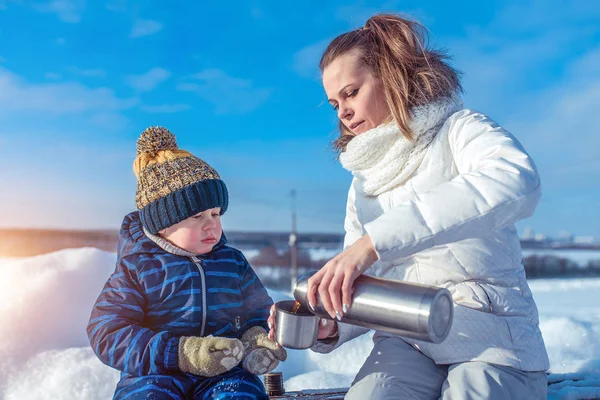 Een vrouw giet een warme drank van een thermos in een mok thee, haar kind, haar jongen zoon is 3 jaar oud, in de winter buiten in het weekend vakantie, met een hapje op de natuur lunch. — Stockfoto