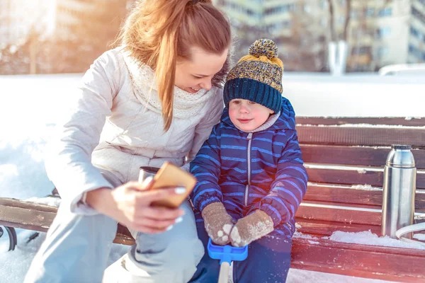 Женщина-мать с ребенком фотографируется на скамейке в городе зимой, в теплой одежде, маленький мальчик 3 лет в синем комбинезоне и шляпе. В магазине термос с горячим напитком . — стоковое фото