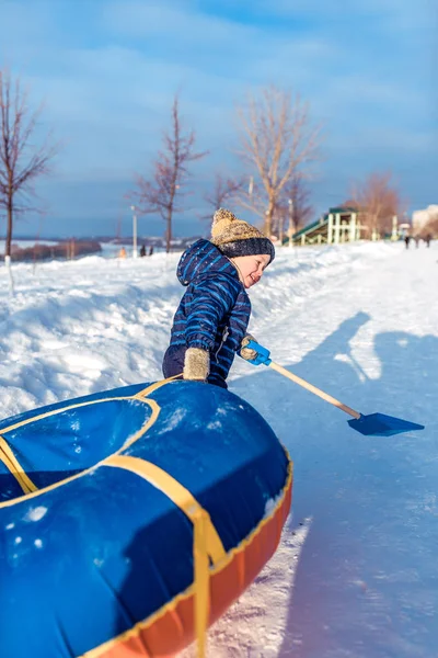 Ένα μικρό αγόρι 3 ετών, το χειμώνα στο δρόμο, σέρνει σωλήνες, για ιππασία από ένα λόφο, στα χέρια ενός παιδικού κουπί, παίζοντας, διασκεδώντας, χαλαρώνοντας τα Σαββατοκύριακα, χιονοπτώσεις στο παρασκήνιο. — Φωτογραφία Αρχείου