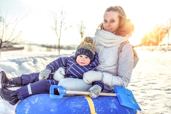 Schöne Mutter mit Baby 3 Jahre alt, sitzend aufblasbare Schläuche, vom Hügel herunter rollen, ausruhen, Winter-Open-Air-Wochenende im Ferienort, im Hintergrund Schneeverwehungen. — Stockfoto