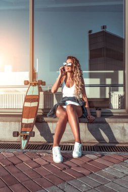 Beautiful girl summer city calls on phone, skate longboard. Happy smiling. Concept fashion style, trends of youth, modern idea clothing and leisure. Tanned skin has long hair. Emotions joy fun. clipart