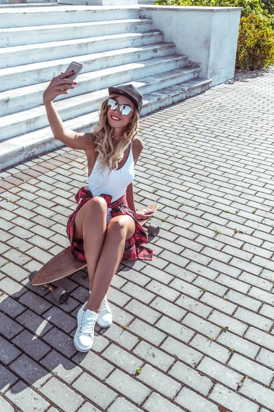 Красива жінка літнього міста фотографує на телефоні біле тіло, дошку, скейтборд. Щасливий усміхнений. Концепція стилю моди, тенденції молоді, сучасна ідея одягу та дозвілля . — стокове фото
