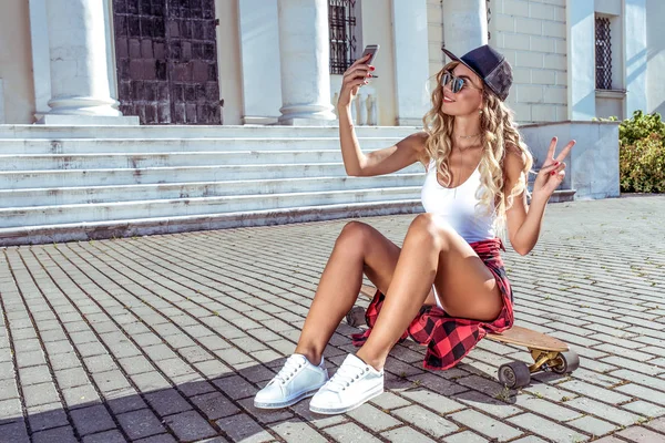 Beautiful girl in the summer city takes pictures on phone, free space, skate longboard. Happy smiling. The concept of fashion style, trends of youth, modern idea of clothing and leisure. — Stock Photo, Image