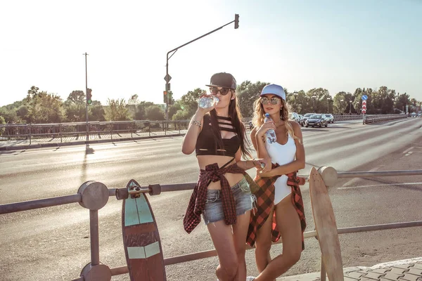 To kvinder veninder, stå vejkryds, skate, drikke vand fra flaske, longboard, ledig plads baggrund vej asfalt bil. Koncept mode stil tendenser ungdom, moderne ide tøj og fritid - Stock-foto