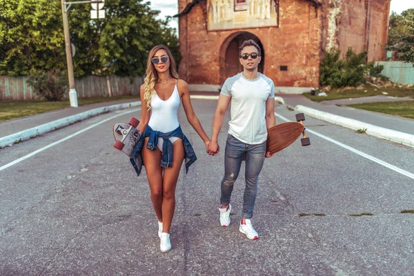 Pasangan muda berjalan di kota musim panas, skate longboard. Gaya mode remaja, tren rekreasi modern, figur olahraga kecokelatan. Emosi kebahagiaan, kesenangan, relaksasi, memiliki akhir pekan yang menyenangkan . — Stok Foto