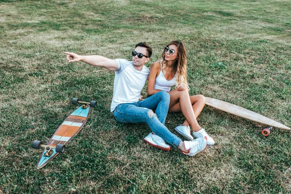 Junges Paar Mann Frau, sitzen auf Gras Sommerstadt, Skate Longboard. Jugendmode, moderner Freizeittrend, gebräunte Sportfigur. Emotionen campen im Urlaub, Spaß haben am Wochenende. — Stockfoto
