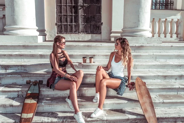 Zwei Freundinnen, Sommerstadt, Skateboards, Longboard-Tassen Kaffeetee. Sie reden entspannt mit der braungebrannten Gestalt. Hintergrund Schritte Gebäude Spalte. Modestil, moderner Lebensstil. — Stockfoto