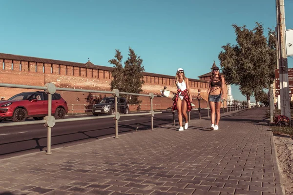 Zwei Studentinnen gehen durch die Sommerstadt, Skateboards, Longboard-Badeanzüge. ruhende lange Haare braun gebrannte Figur. Hintergrund Straßenautos. Modestil, moderner Lebensstil. Freiraum für Text. — Stockfoto